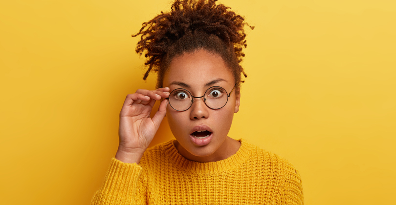 Mulher negra de óculos, com expressão se surpresa, vestindo camiseta amarela sob um fundo amarelo.