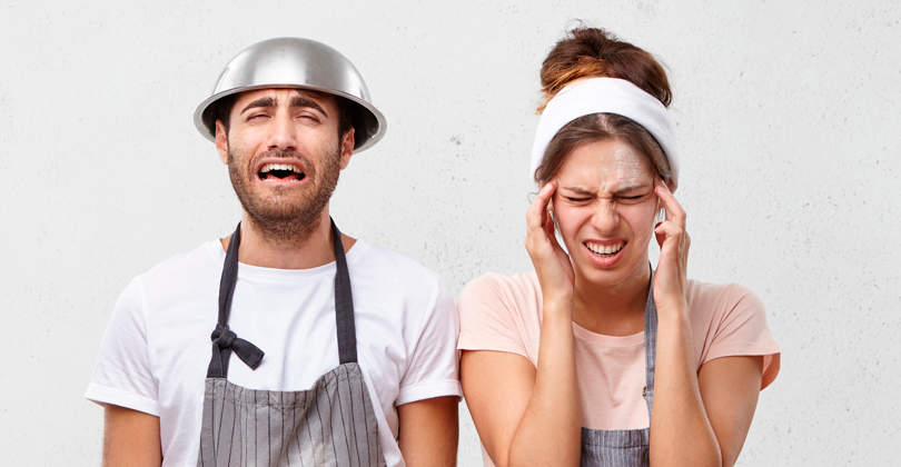 Pizzaiolos preocupados com a dificuldade de porcionar os insumos. Homem com a bacia na cabeça e mulher com as mãos indicando dor de cabeça. Ambos com a expressão de estresse.